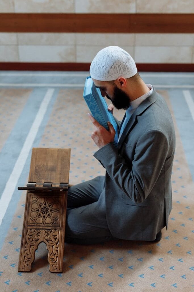 pexels-photo-8489307 A bearded Muslim man in a suit kneels indoors in prayer, holding the Quran on a wooden stand.